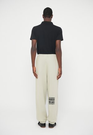 Άνδρας που στέκεται με πλάτη, φορώντας μαύρο πόλο μπλουζάκι, ανοιχτό γκρι φόρμες με την ένδειξη "Hardcore Wellness Berlin 2025-2027" στο αριστερό πόδι, και μαύρα παπούτσια.