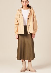 Giacca impermeabile beige con cappuccio e polsini elastici, abbinata a una gonna midi verde. Stivaletti beige chiaro con una suola robusta.