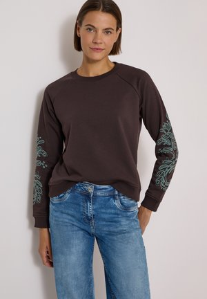 Femme portant un sweat-shirt marron foncé avec des motifs floraux verts sur les manches et un jean bleu, debout contre un mur clair.
