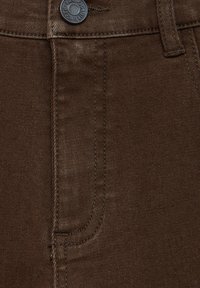 Gros plan d’un tissu denim marron montrant une poche avant et un bouton métallique avec le texte « Pacific Republic ».