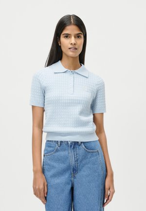 Kvinne med langt mørkt hår som har på seg en lyseblå kortermet teksturert poloskjorte og høytlivsblå jeans, stående mot en ensfarget bakgrunn.