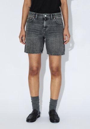 Sorte denimshorts med lige snit, der har fem lommer, synlige syninger og en rå kant. Bæres med grå sokker og sorte loafers.