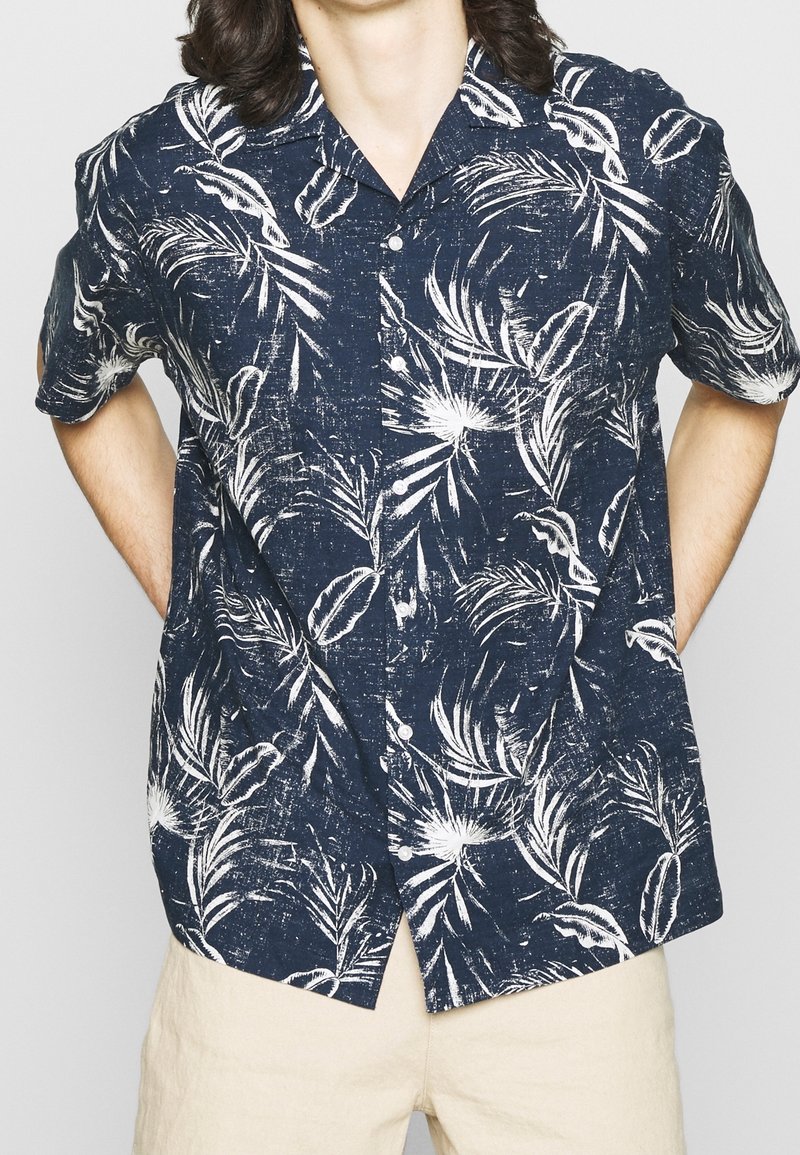 Navy short-sleeve button-up shirt with a white tropical leaf print; lightweight fabric with a textured finish, featuring a classic collar.
