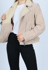 Frau in beiger Wildlederjacke mit cremefarbener Schafsfellverkleidung, weißem Hemd und schwarzen Hosen, die die Jacke geöffnet hält, um das Futter zu zeigen.