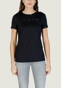 Vrouw draagt een zwart Armani T-shirt met gestikte logo, gecombineerd met lichtgrijze jeans en rode nagellak, staand tegen een effen achtergrond.