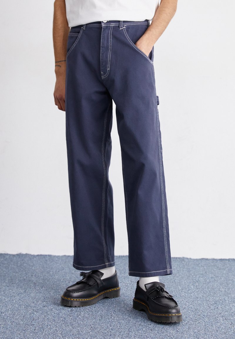Personne portant un pantalon large bleu marine avec des surpiqûres contrastantes, des mocassins noirs à pampilles et des chaussettes blanches, debout sur un tapis bleu contre un mur blanc.
