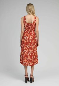 Robe maxi en rouge rouille avec imprimé floral blanc, dotée d'un dos carré, d'une taille smockée et de larges bretelles. Associée à des sandales à talons blocs marron.
