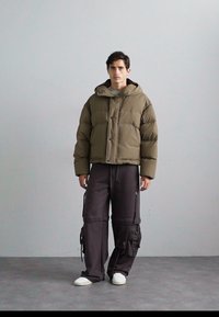 Khaki-Steppjacke mit Kapuze, mit gesteppten Abschnitten und vorderen Knöpfen, kombiniert mit lockeren, dunkelgrauen Cargo-Hosen und weißen Sneakers.