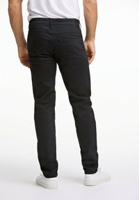 Zwarte jeans met een slim fit, voorzien van achterzakken en een gestructureerde stof. Gecombineerd met witte sneakers. Zicht van achteren.