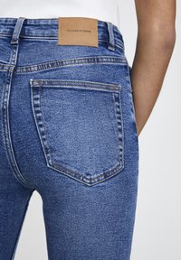 Los pantalones vaqueros de mezclilla azul presentan un diseño típico de cinco bolsillos, costuras visibles y un parche de cuero marrón con la marca en la cintura.