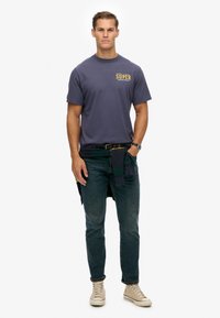 Navyblauwe katoenen t-shirt met "SUPER" in gele tekst, gecombineerd met donkere jeans en beige hoge sneakers. Groene geruite shirt geknoopt om de taille.