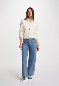 Krämfärgad stickad cardigan med rund hals, knäppning framtill och ribbade manschetter, kombinerad med högmidjade blå jeans och vita sneakers.