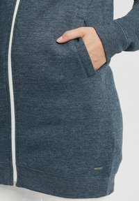 Mörkblå zip-up hoodie av mjukt tyg, med sidofickor, en vit dragkedja och en liten logotag nära fållen.