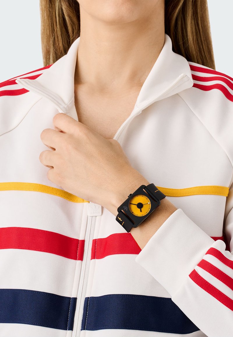 Montre rectangulaire noire avec un cadran jaune, portée au poignet. La veste blanche a des rayures horizontales rouges, jaunes et bleues.