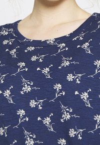 Blue short-sleeve top with a round neckline, featuring a small white floral pattern on a textured fabric. Subtle, casual design detail.