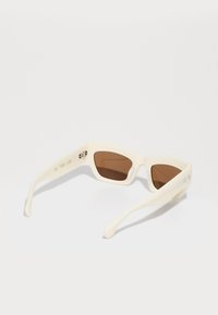 Lunettes de soleil de couleur crème avec un design géométrique, verres marron et branches fines et courbées. Présentent des éléments en métal argenté sur la charnière.