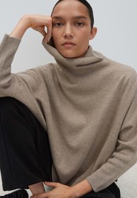 Frau mit dunklem Haar, das zurückgebunden ist, trägt einen beigen Rollkragenpullover und schwarze Hose. Sie stützt ihren Kopf auf die Hand und sitzt vor einem schlichten Hintergrund.