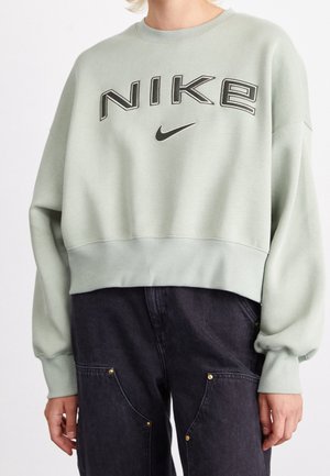 Osoba nosząca jasnozieloną bluzę Nike z czarnym logo oraz ciemne, wysokie spodnie z dużymi przodem kieszeniami i złotymi nitami.