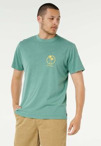 Uomo che indossa una maglietta verde acqua con logo di palma gialla e pantaloni di velluto a coste color beige, che guarda di lato su uno sfondo bianco semplice.