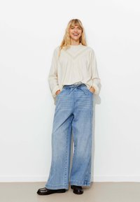 Pull en maille beige clair avec un design texturé, associé à un jean en denim bleu clair à jambes larges. Des mocassins noirs à bout carré complètent l'ensemble.