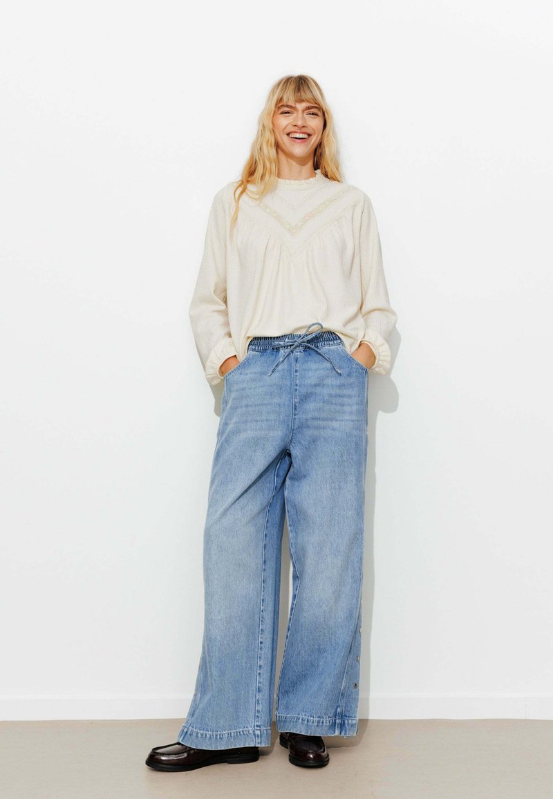 Pull en maille beige clair avec un design texturé, associé à un jean en denim bleu clair à jambes larges. Des mocassins noirs à bout carré complètent l'ensemble.