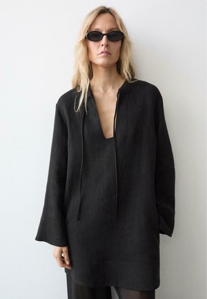 Donna con lunghi capelli biondi che indossa piccoli occhiali da sole neri e una tunica nera larga con scollo a V profondo su uno sfondo chiaro e uniforme.