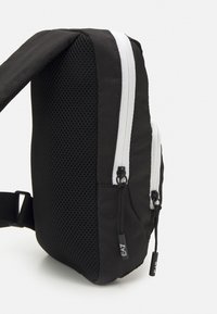 Zaino nero con un lato in rete testurizzata, doppie cerniere bianche e tirazip neri; presenta una sola tracolla regolabile e un design elegante.