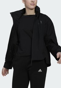 Zwarte waterdichte jas met een hoge kraag en rits, gelaagd over een getailleerd zwart shirt en zwarte broek met een wit Adidas-logo.