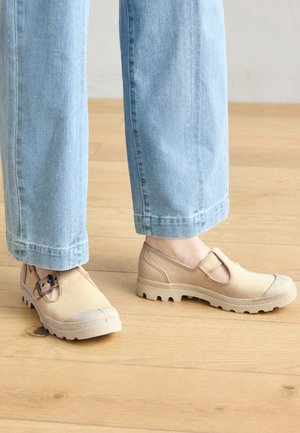 Chaussures slip-on beiges avec une semelle en caoutchouc, présentant une sangle et un détail de boucle, associées à un jean bleu clair sur un sol en bois.