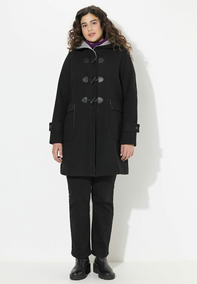 Cappotto nero di lana con chiusure a toggles, grandi tasche anteriori e fodera in tessuto grigio per il cappuccio. Sotto indossa un pantalone nero e stivaletti.