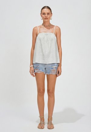 Mujer con camiseta sin mangas blanca, shorts de mezclilla azul claro desgastados y sandalias con cordones y cuentas, de pie sobre fondo blanco.