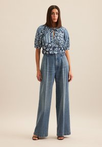 Blusa blu con motivo floreale, maniche a sbuffo, scollo a chiave, abbinata a jeans in denim chiaro a vita alta e gamba larga. Sandali neri.