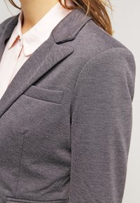Blazer gris ajusté avec une texture lisse, des revers crantés et une poche poitrine unique, associé à une chemise rose clair à col en dessous.