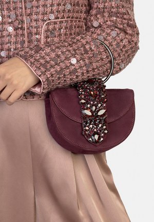 Runde, bordeauxrote Wildlederhandtasche mit einer klappbaren Verschlusslasche, die rote und klare Kristalle als Schmuck hat. Wird in der Hand getragen, kombiniert mit einem pinken Outfit.