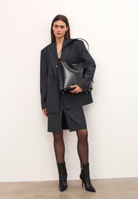 Mujer con un blazer de rayas oversized y falda, sosteniendo un gran bolso de cuero negro, vistiendo medias negras transparentes y botas de tacón puntiagudas.
