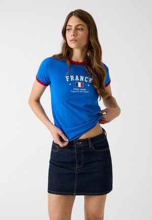 Jeune femme ajustant un t-shirt bleu "France" avec un liseré rouge, associé à une jupe en jean foncé, debout devant un fond blanc.