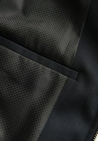 Textil negro con patrón hexagonal texturizado; presenta un panel de tela suave y un ribete negro horizontal. Detalle de cremallera en la parte inferior derecha.