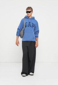 Joven con sudadera azul de GAP, pantalones anchos negros, bolso bandolera gris, grandes gafas de sol negras, de pie frente a un fondo blanco.