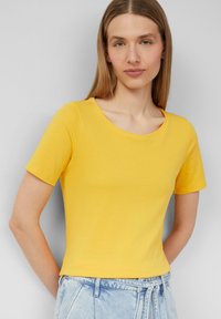 T-shirt jaune à manches courtes avec un col rond, fabriquée en tissu coton lisse. Associée à un short en denim bleu clair taille haute.