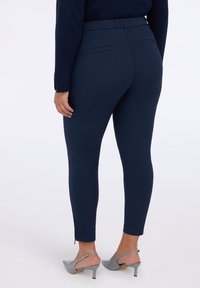 Marineblaue enganliegende Hosen mit einem elastischen Bund, Reißverschlussmanschetten und zwei Gesäßtaschen. Das Material wirkt glatt und elastisch.