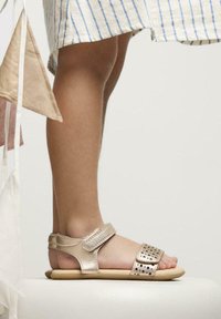 Sandalias metálicas doradas con tiras de velcro, detalles perforados y una suela beige. El diseño ofrece un estilo de punta abierta para una mejor ventilación.