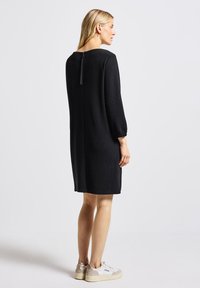 Robe en tricot noir avec un col rond et des manches bouffantes longues. Détail de fermeture éclair au dos et coupe décontractée, associée à des baskets blanches et beiges.