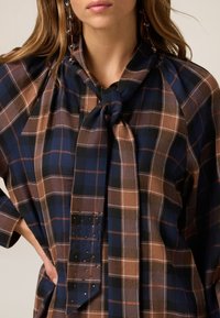 Skjorte i tern i blå, brun og sort stof med bundet hals, der har et tekstureret panel med nitter og en løstsiddende pasform.