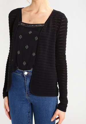Femme portant un cardigan noir à maille ouverte par-dessus un chemisier noir à motifs et un jean bleu taille haute, debout devant un fond blanc.