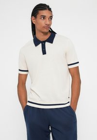 Camisa polo beige claro con cuello y ribetes azul marino, con mangas cortas, botones en el cuello y detalles acanalados.