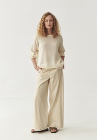 Maglione lavorato a maglia beige chiaro con una vestibilità rilassata e texture a coste, abbinato a pantaloni a gamba larga crema e infradito marroni.