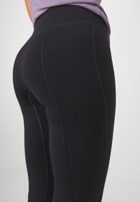 Czarne legginsy o gładkiej fakturze, z kontrastowymi szwami i dopasowanym krojem, pokazane z boku.