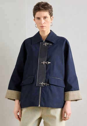 Tmavomodrá bunda s veľkým golierom, zapínaním na zips a kovovými sponami. Má dve vrecká vpredu a manžety s kockovaným vzorom pre kontrast.