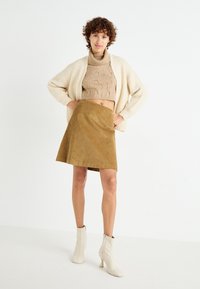 Maglione corto lavorato a maglia beige, cardigan aperto crema e un gonna a campana in corduroy marrone. Il modello indossa stivaletti bianchi alla caviglia con tacco.