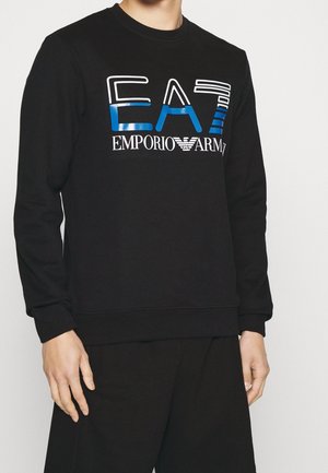 Osoba ubrana w czarną bluzę z długim rękawem z niebiesko-białym logo "EA7 Emporio Armani" na piersi, w połączeniu z czarnymi spodniami.